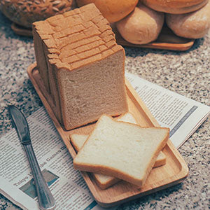 A loaf of white bread is cut into two slices, one of which is placed on a wooden board next to a knife, with more slices stacked in the background.