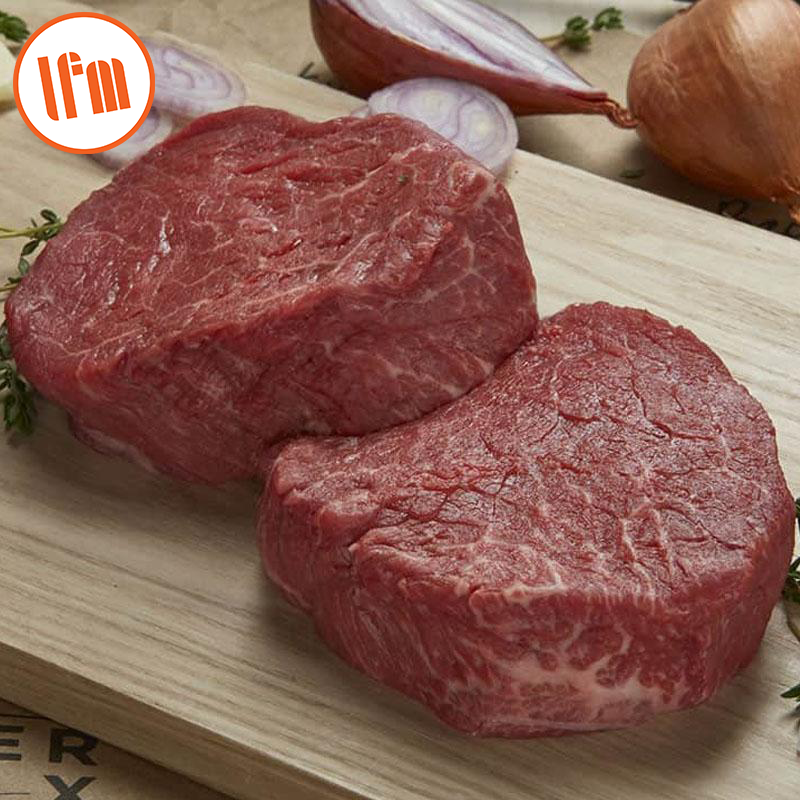 Two raw beef tenderloin steaks on a wooden cutting board with a knife and onion slices in the background.