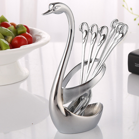 A silver-colored stainless steel swan-shaped holder for fruit forks and spoons, placed on a white surface with a bowl of fruit in the background.